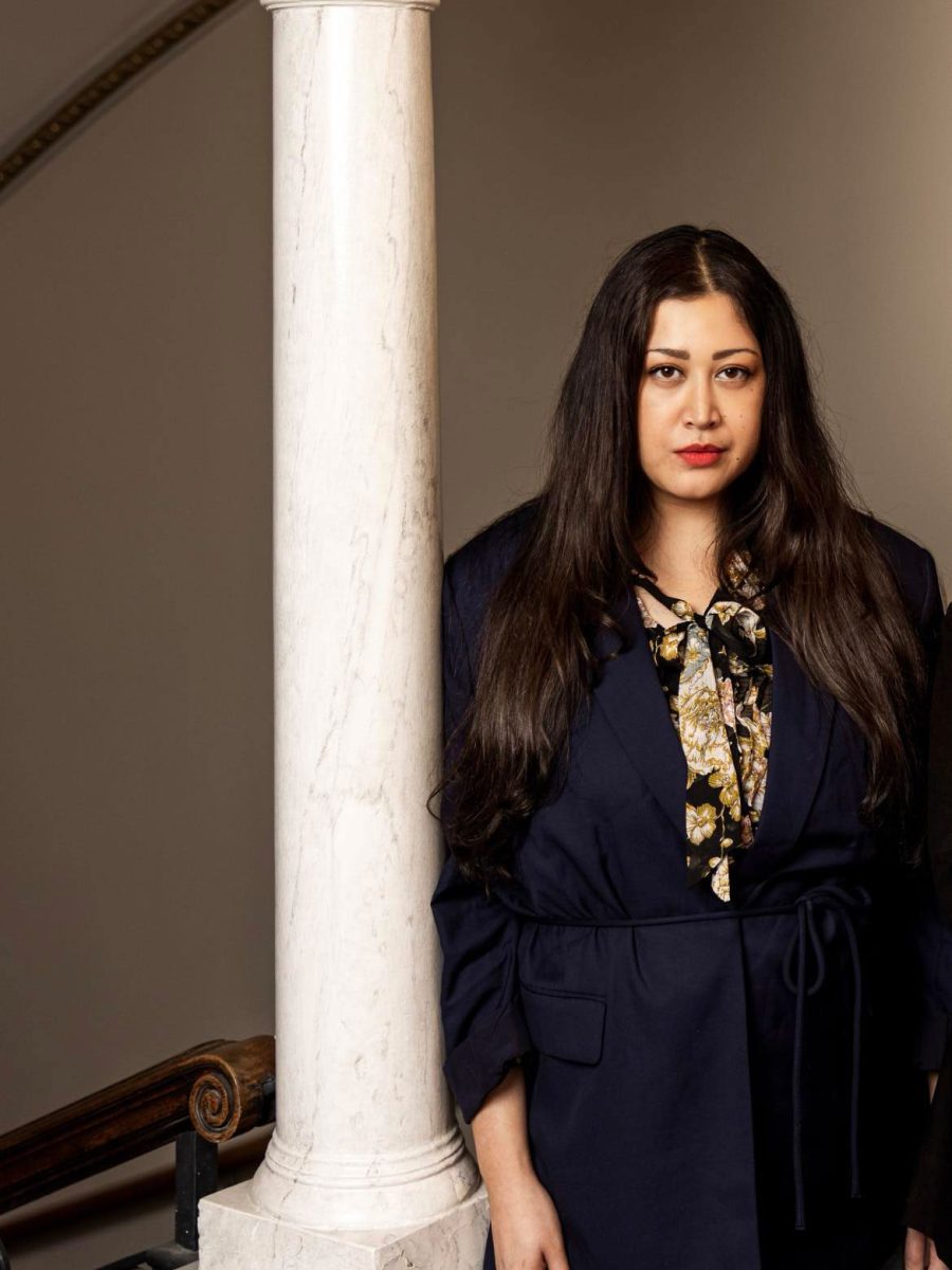 Louise Neo, grundare av Sistjejer, med svart hår och rött läppstift står lutat mot en pelare i svart kavaj.